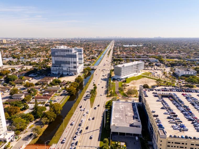Blick auf eine belebte Straße vor dem Campus der FIU in Miami mit Palmen und vorbeiziehenden Autos im Vordergrund.