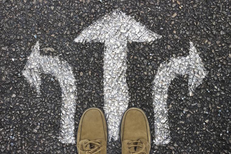Perspektive von Schuhen auf asphaltiertem Boden mit drei Richtungsweisern, die symbolisch für Entscheidungsfindung in der Planungsphase stehen.