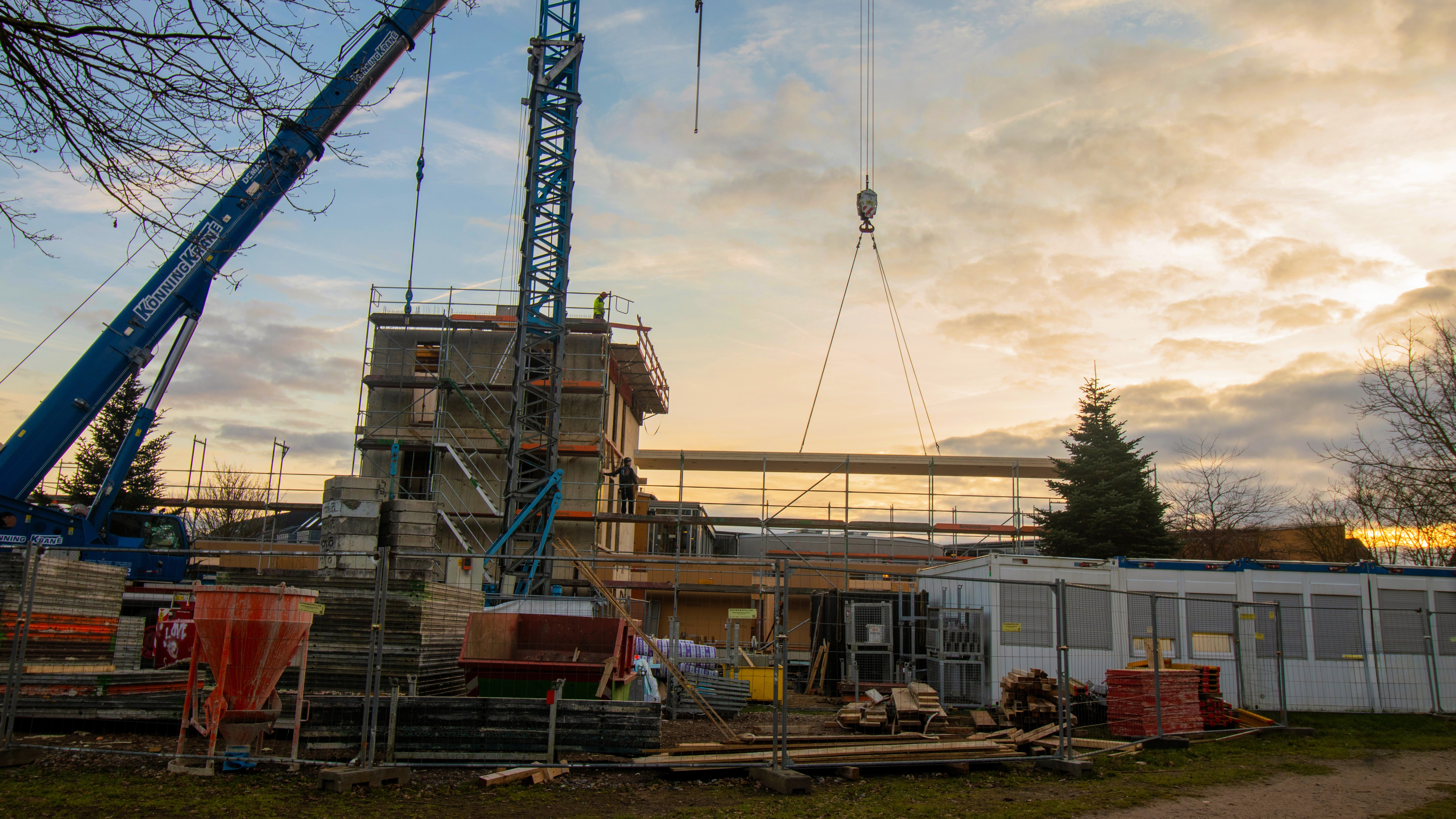 Baustelle einer Grundschule im Rohbaustadium: Mobilkran und Turmdrehkran heben lange Holzbauelemente, Gerüst und Baucontainer auf der Baustelle