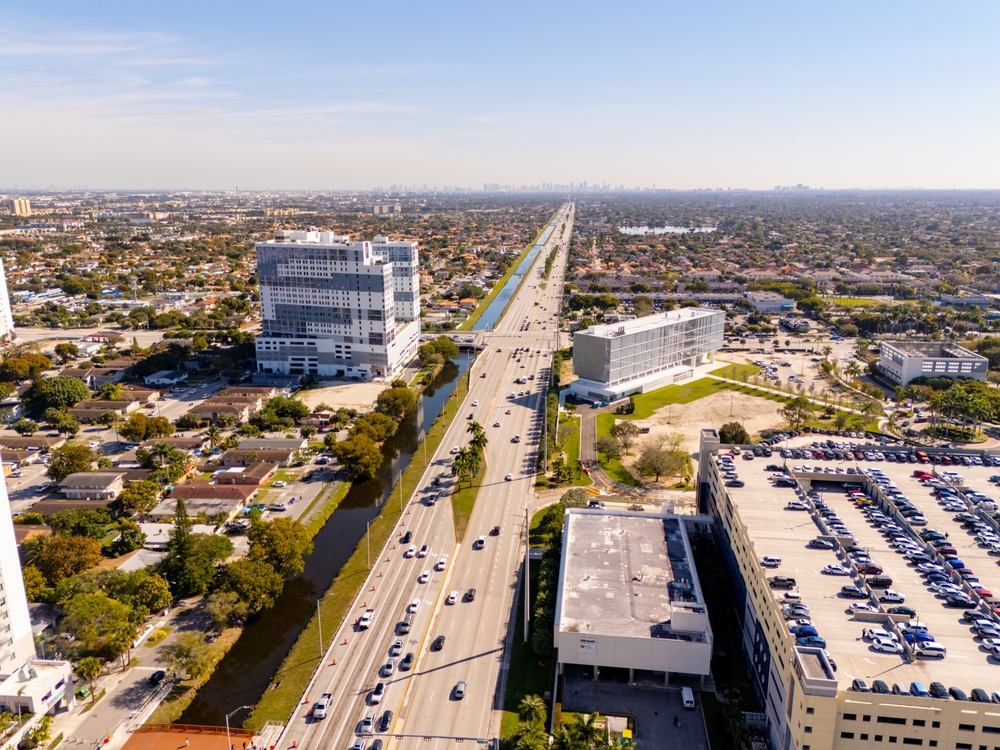 Vue d’une Route animée devant le Campus de la FIU à Miami avec des palmiers et des voitures qui passent au premier plan.