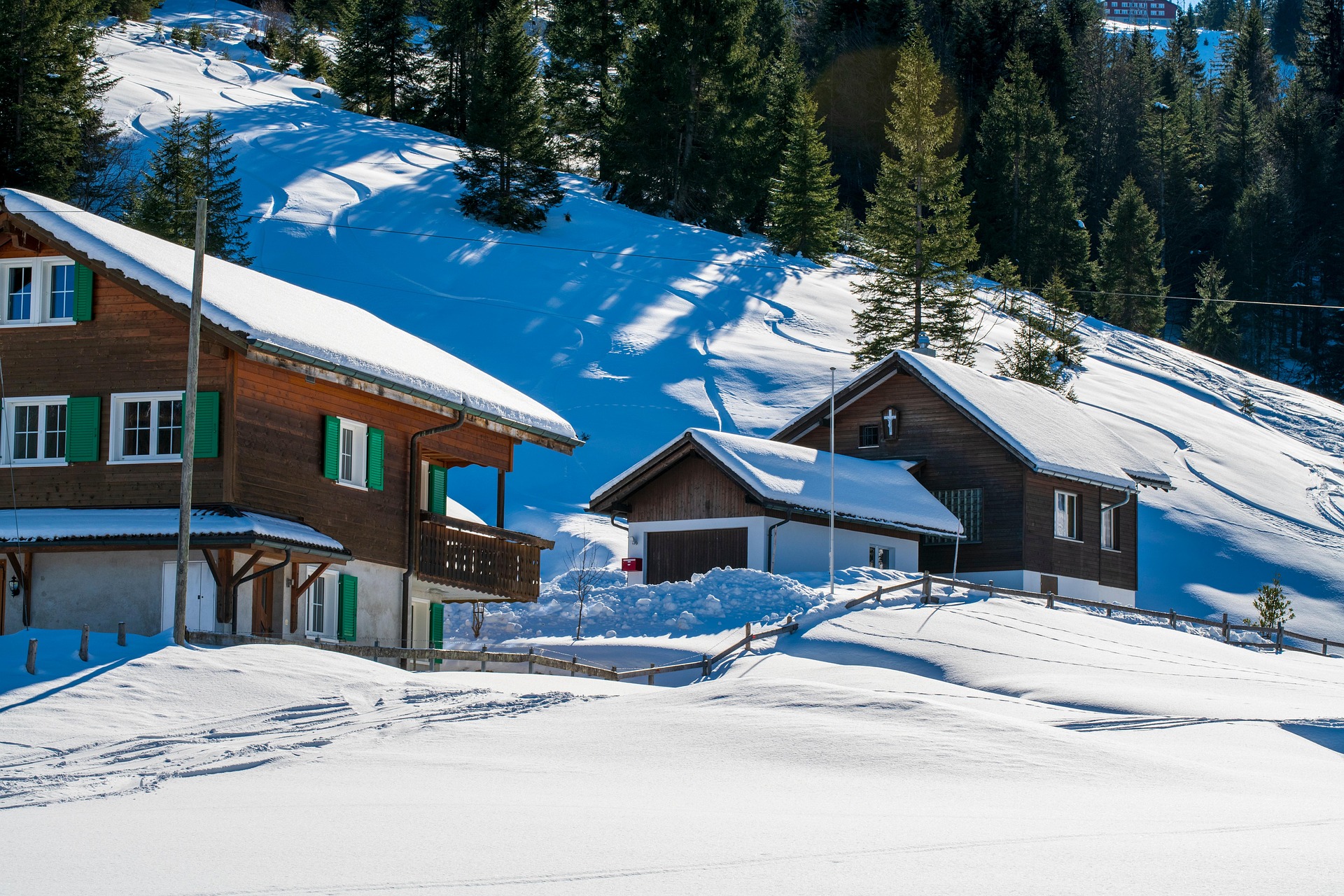 Deux maisons traditionnelles en bois aux volets verts et aux toits enneigés dans un paysage alpin hivernal par temps clair et ensoleillé.