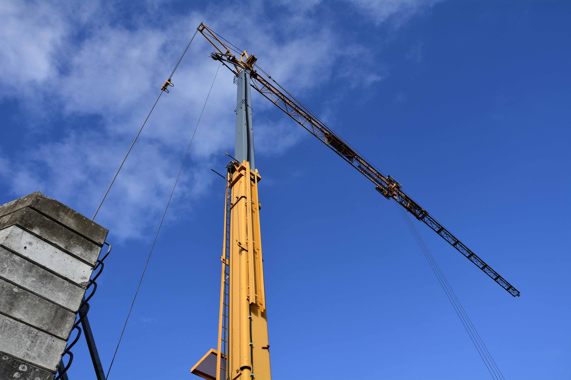 Gru gialla con braccio e gancio di carico davanti a un cielo blu, posizionata accanto a una parete di cemento in un cantiere.