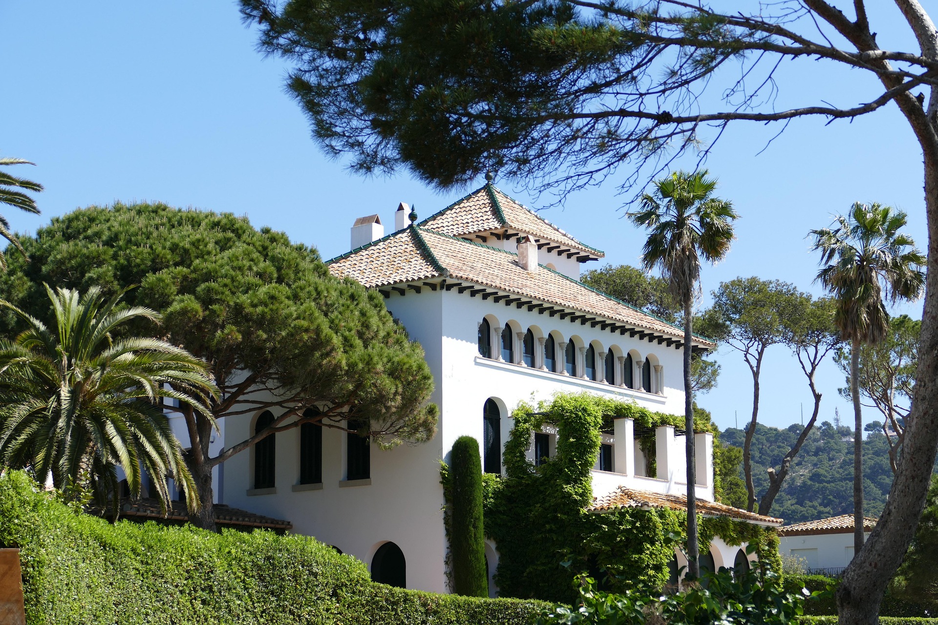 Mediterrane Villa mit weißer Fachada, Ziegeldach und Arkadenfenstern, umgeben von Pinien und Palmen in sonniger Landschaft der Costa Brava.