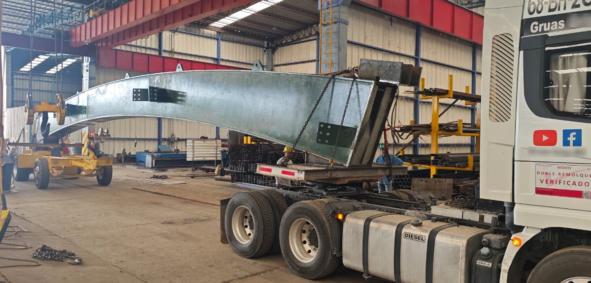 Viga em arco de aço para ponte sendo içada e carregada com guindastes sobre caminhão de transporte em oficina de fabricação metálica.