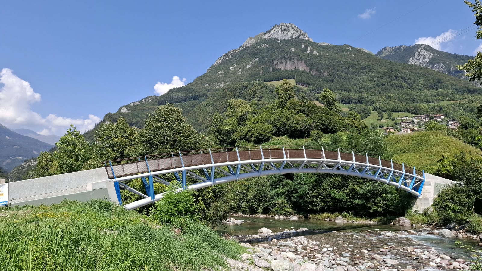 意大利高山背景前跨越河流的钢制弧形行人桥